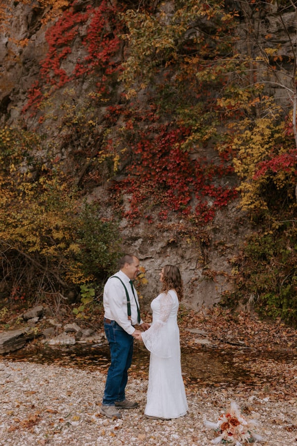 A couple holds hands looking at each other while they pose for a photo in front of a bluffs with vibrant fall foliage during their autumn elopement at Steel Creek Campground on the Buffalo National River in Arkansas.