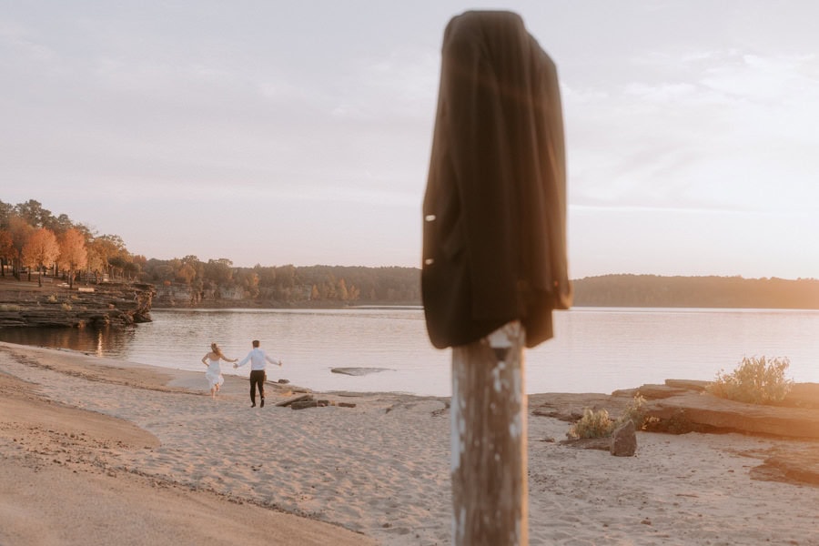 A groom playfully chases after his bride on a beach in Heber Springs, Arkansas during their adventure elopement.