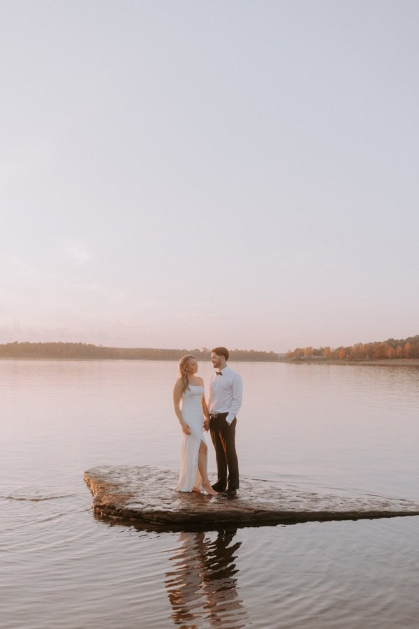A couple holds hands on a rock low in the water of Greers Ferry Lake while taking elopement photos in Heber Springs, Arkansas.