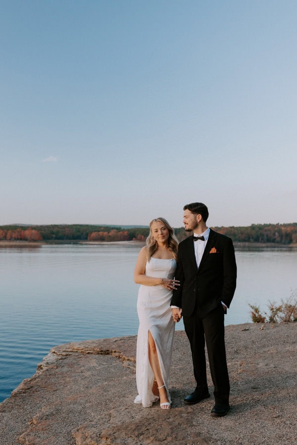 A couple holds hands on a bluff overlooking a vibrant blue Greers Ferry Lake while taking elopement photos in Heber Springs, Arkansas.