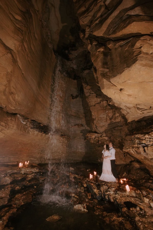 A couple stands embracing inside of a cave looking up at a waterfall during their Arkansas adventure elopement at Lost Valley Trail in Ponca.