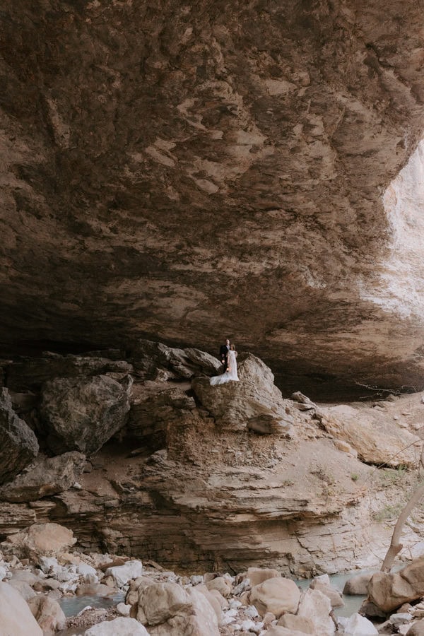 A couple stands on a rock high off the ground inside of a large cave while exploring during their Arkansas elopement.