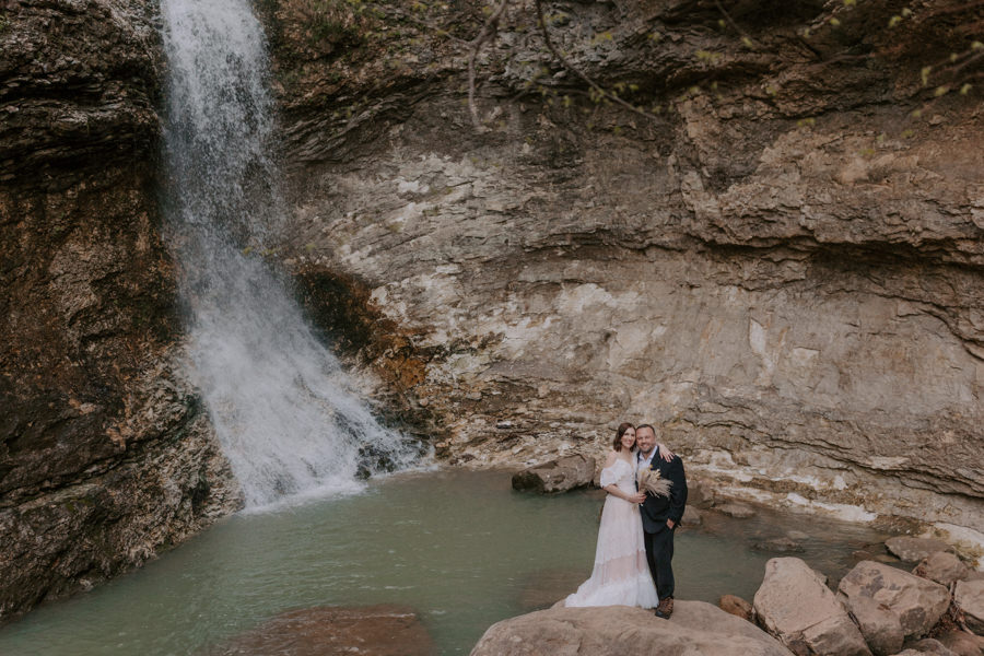 a couple embraces each other and smiles for the camera for a photo in front of a waterfall during their adventure elopement in Ponca, Arkansas at Lost Valley Trail.