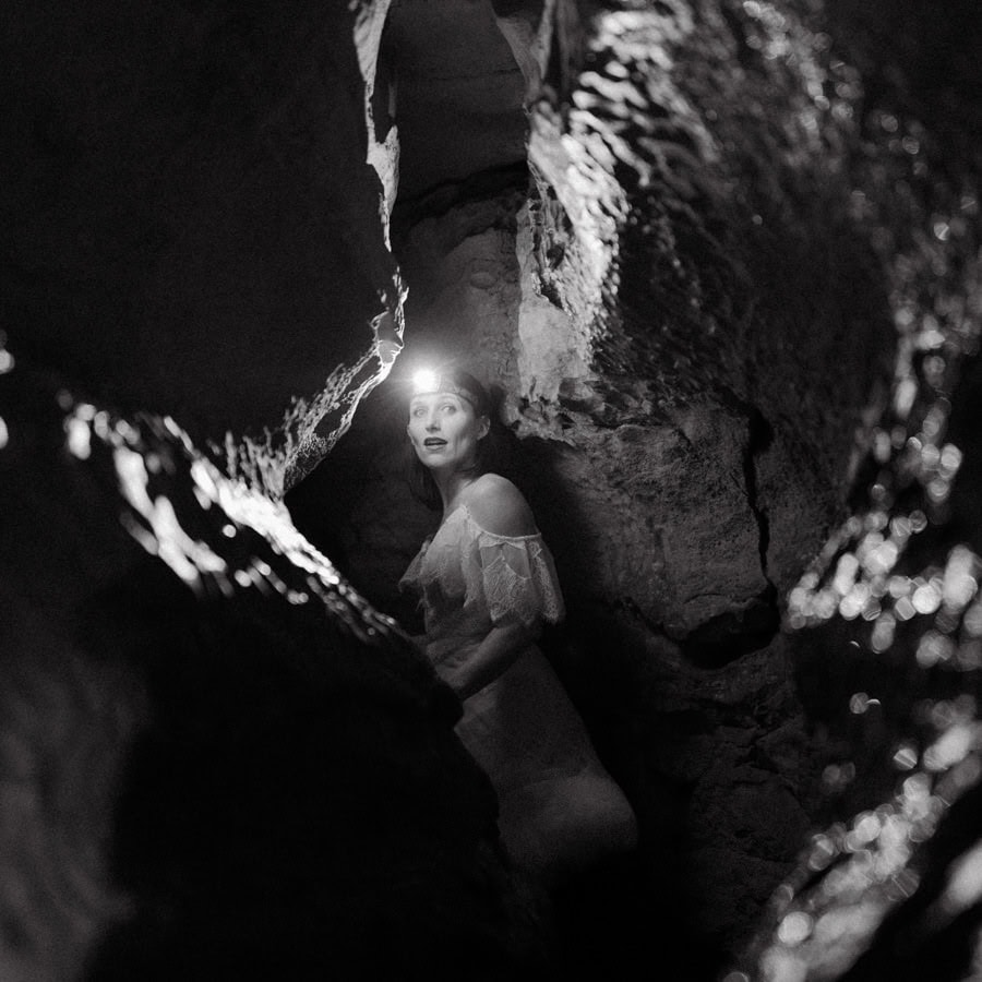 a bride looks back at the camera with a headlamp lighting a narrow cavern while caving during an adventure elopement in Arkansas.
