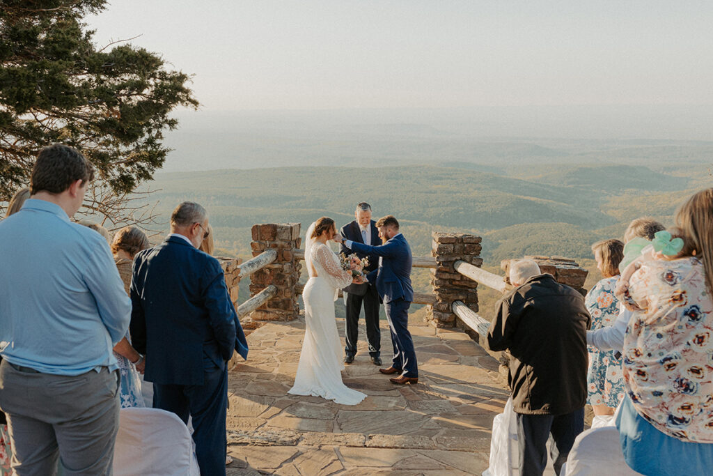 A couple exchange vows in front of their family at Mt. Magazine State Park in Arkansas. The groom is brushing a tear off of the bride's cheek.