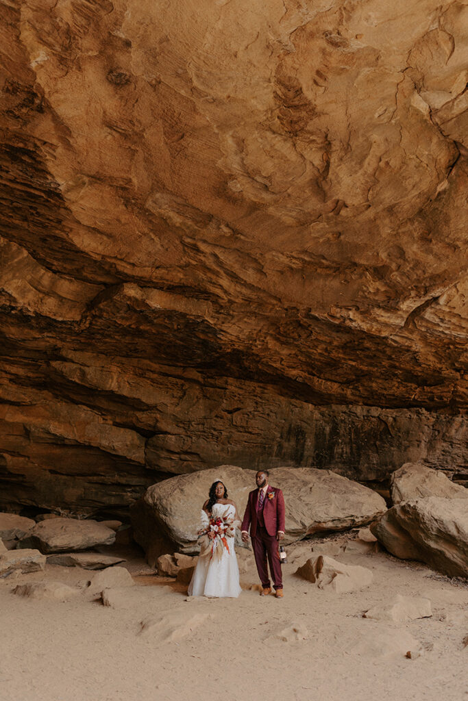 A bride and groom in wedding attire stand inside of the rock house cave at Petit Jean State Park while exploring during their Arkansas adventure elopement.