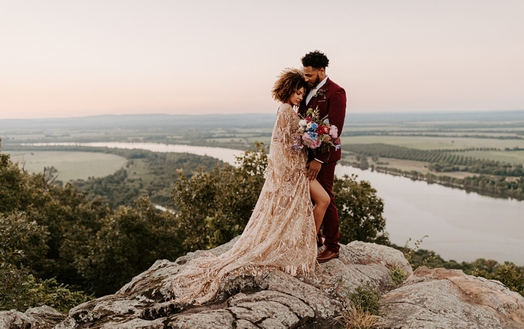 A bride and groom in wedding attire stand on Petit Jean Mountain overlooking the Arkansas River for their elopement at Stouts Point.