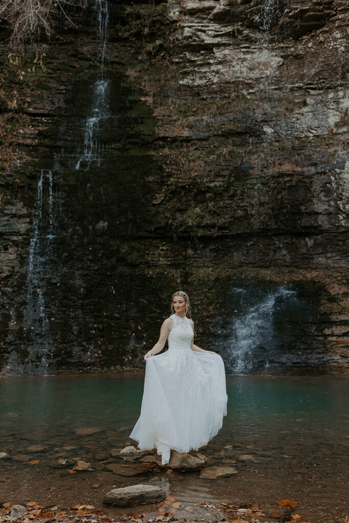 A bride stands on a rock sticking out of a pool of water created by two waterfalls seen behind her during her adventure elopement in Ponca, Arkansas.