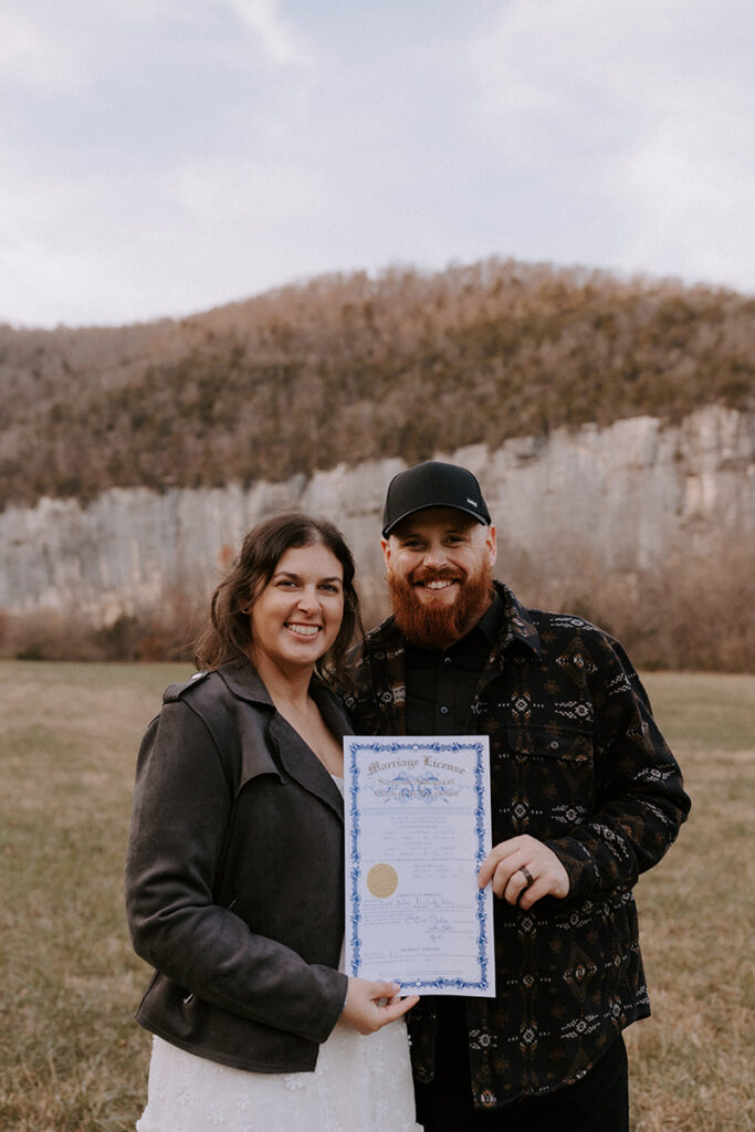 A couple stands in a field at Steel Creek Campground on the Buffalo National River in Arkansas. They're holding their Arkansas marriage license because they just eloped. The naturally painted rocks of Roark Bluff is seen in the background.