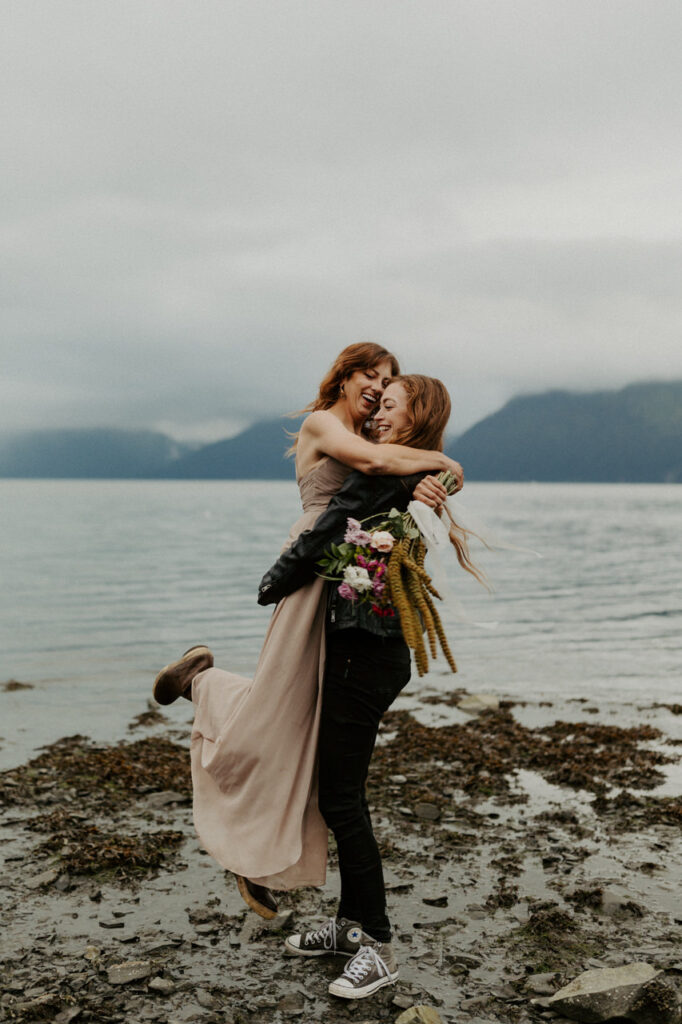 A couple in wedding attire embrace each other while one picks up the other to spin her around while laughing and smiling along a beach in Seward, Alaska during their Kenai Fjords National Park elopement.