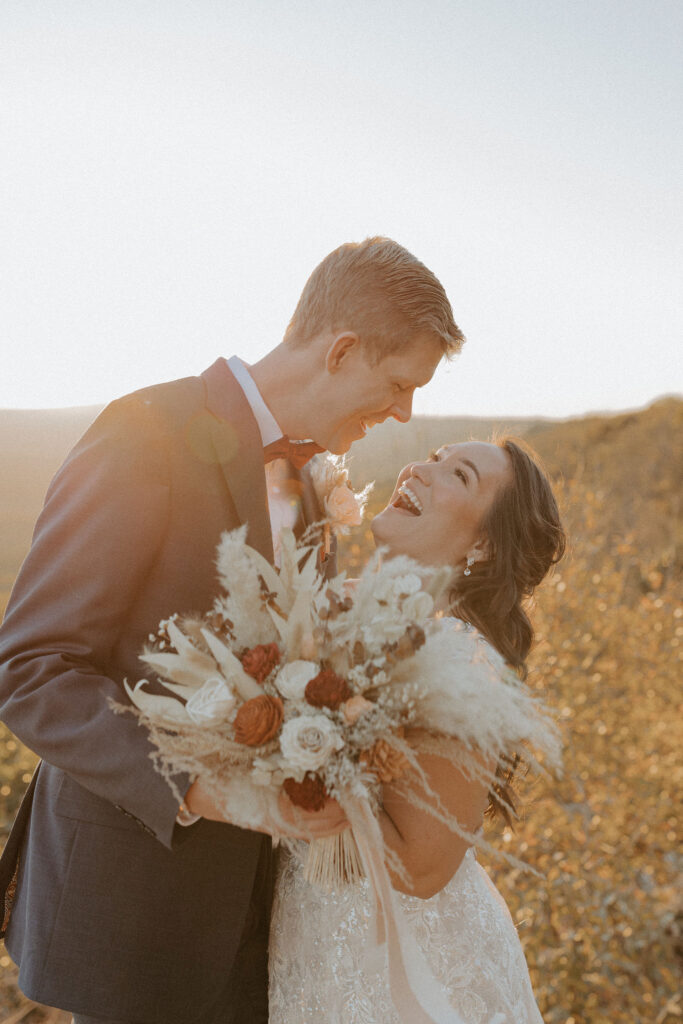 A bride and groom hug each other on a cliffside at Stouts Point on Petit Jean State Park in Arkansas. The bride is looking up at the groom laughing while he holds her dried floral arrangement of white, beige, and red flowers for her.