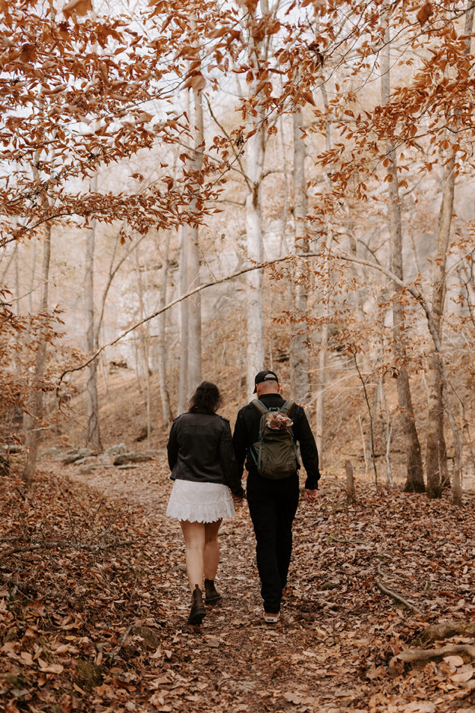 A bride and groom are walking away from the camera down Lost Valley Trail in Arkansas as they head to a scenic spot for their ceremony. It's late fall with many brown leaves on the ground and just a few left in the trees. The groom has a backpack on with the bride's bouquet sticking out of the top.