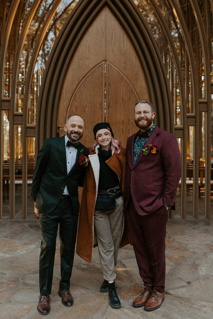 Two grooms stand in front of Cooper Chapel in Northwest Arkansas with their  wedding photographer for a photo together.