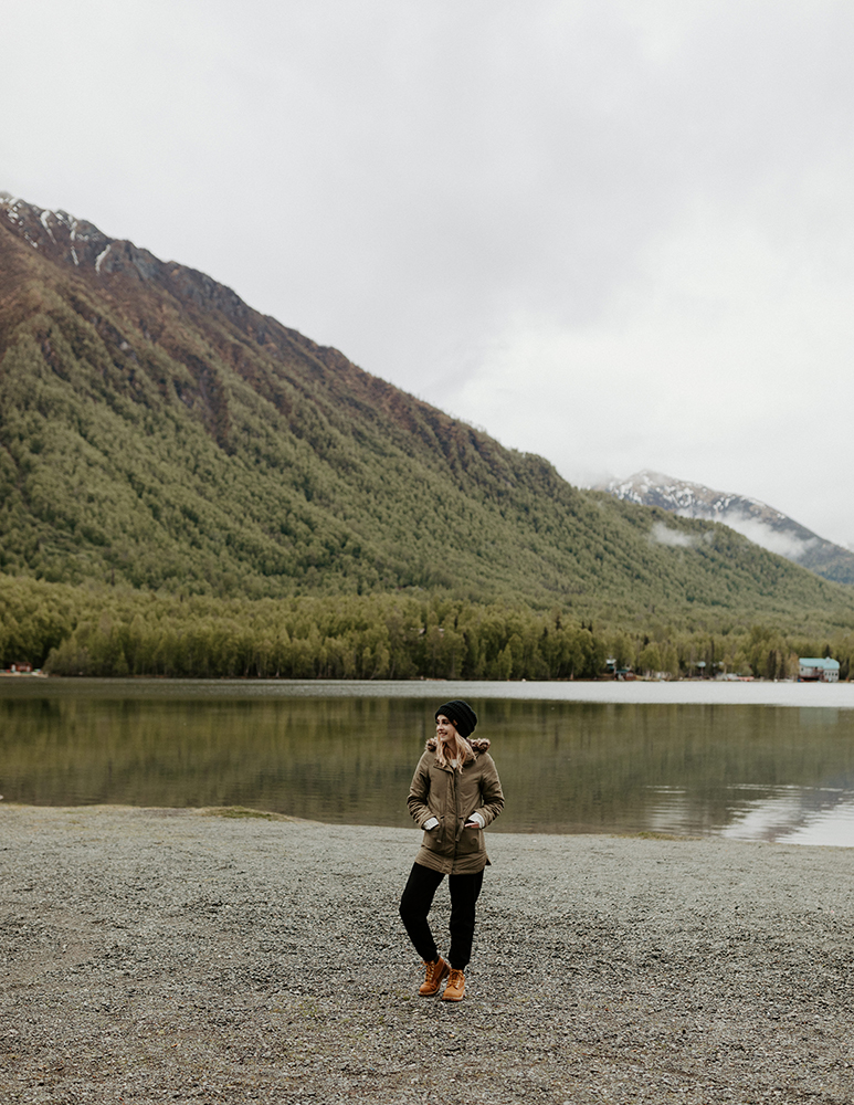 Arkansas + Alaska wedding photographer, Caitlin, stands in front of a cloudy mountain and lake outside of Anchorage, Alaska.