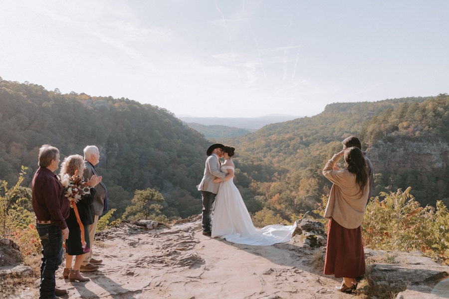A bride and groom kiss during their ceremony surrounded by a few of their relatives and the fall-colored mountains at Mather Lodge Overlook during their Arkansas elopement at Petit Jean State Park.
