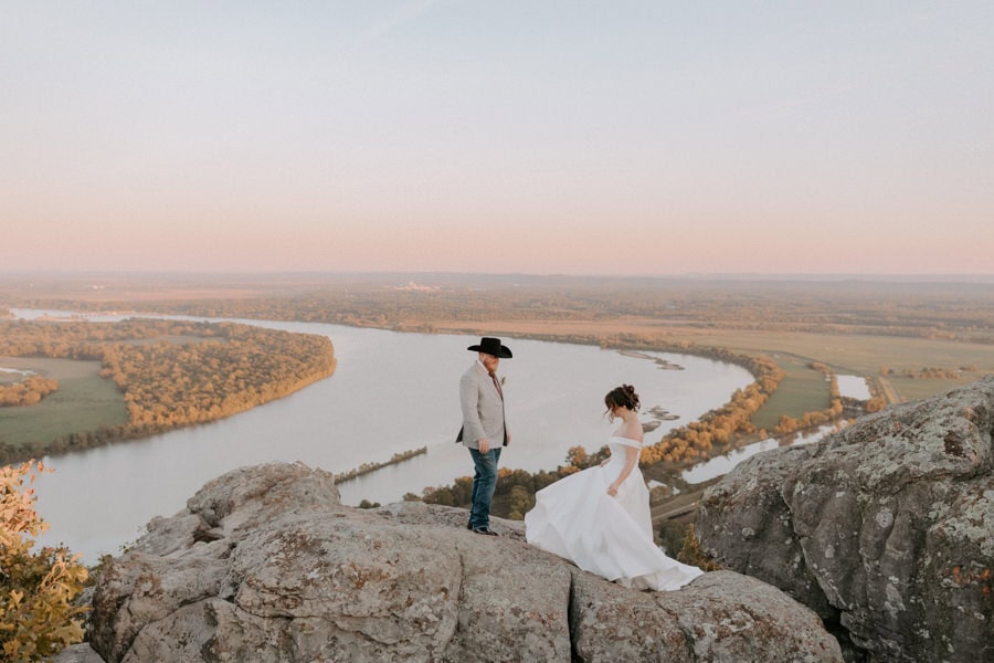 A bride and groom stand on a rocky cliffside overlooking the Arkansas River at Petit Jean State Park. The bride swishes her dress around to show the groom during their elopement in Arkansas.