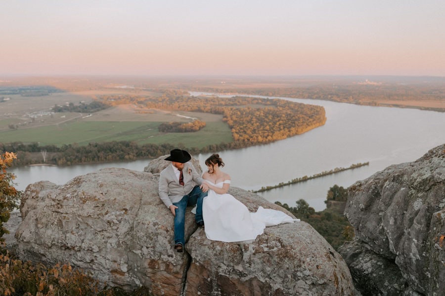A bride and groom sit on a mountaintop with views of the Arkansas River in the background during their Arkansas elopement photos at Petit Jean State Park.