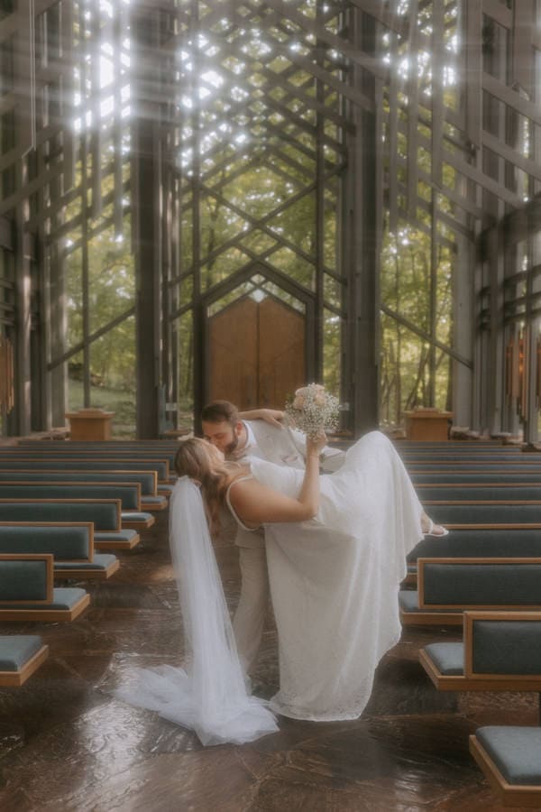 A groom leans his bride down for a dip kiss inside of the glass chapel Thorncrown Chapel in Eureka Springs, Arkansas for their elopement photos.
