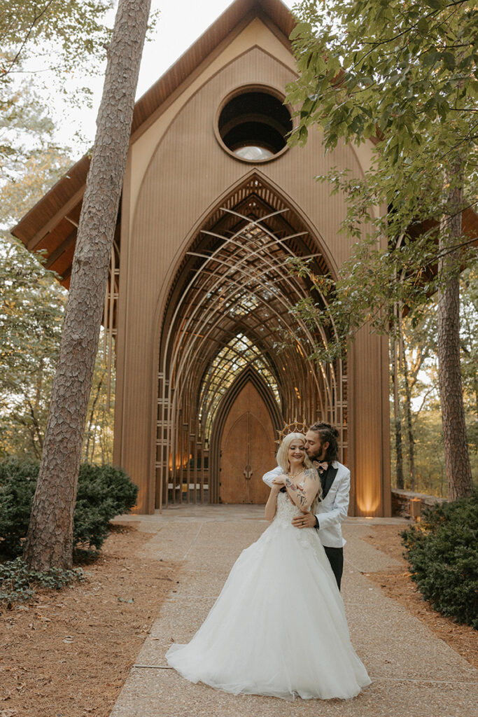 A bride and groom in wedding attire embrace outside of a glass chapel wedding venue in Bella Vista, Arkansas. Cooper Chapel.