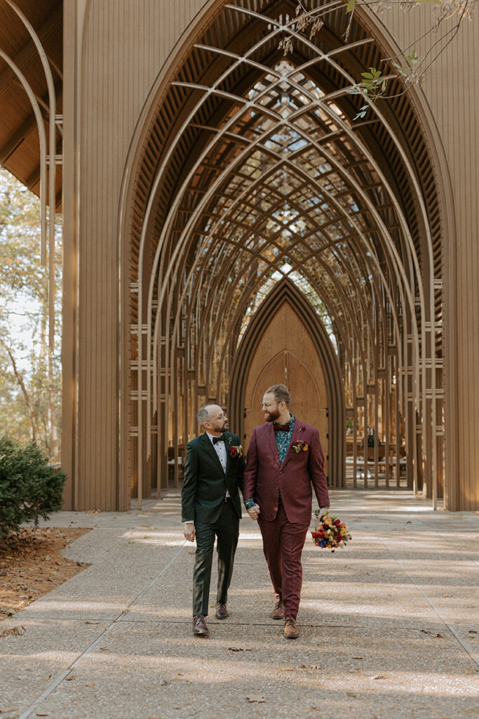 Two grooms walk down a path outside of a glass chapel in Bella Vista, Arkansas after their LGBT wedding. Cooper Chapel wedding venue. 