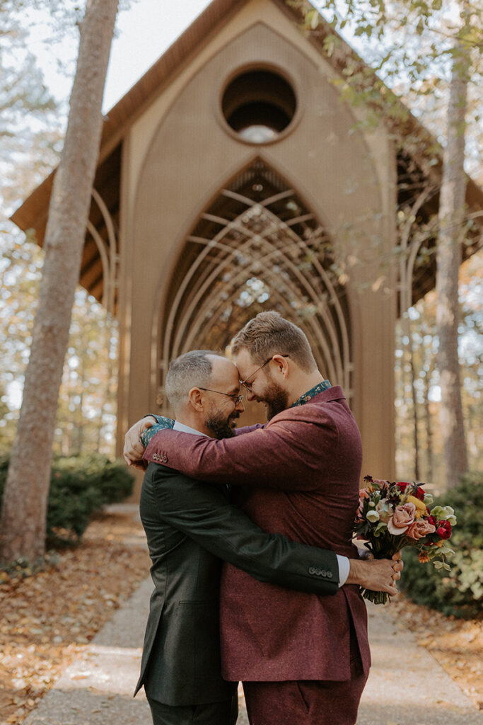 Two grooms in wedding attire embrace outside of their glass chapel wedding venue, Cooper Chapel, in Bella Vista, Arkansas.