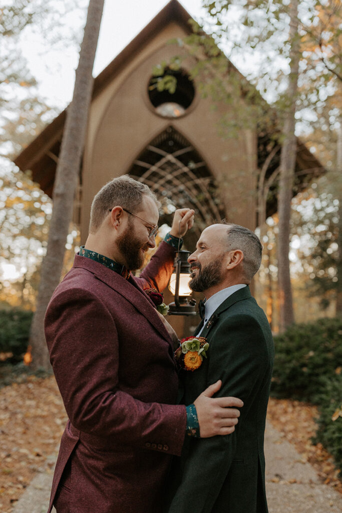 Two grooms stand embracing and smiling outside of their wedding venue, Cooper Chapel in Bella Vista, Arkansas after their elopement.