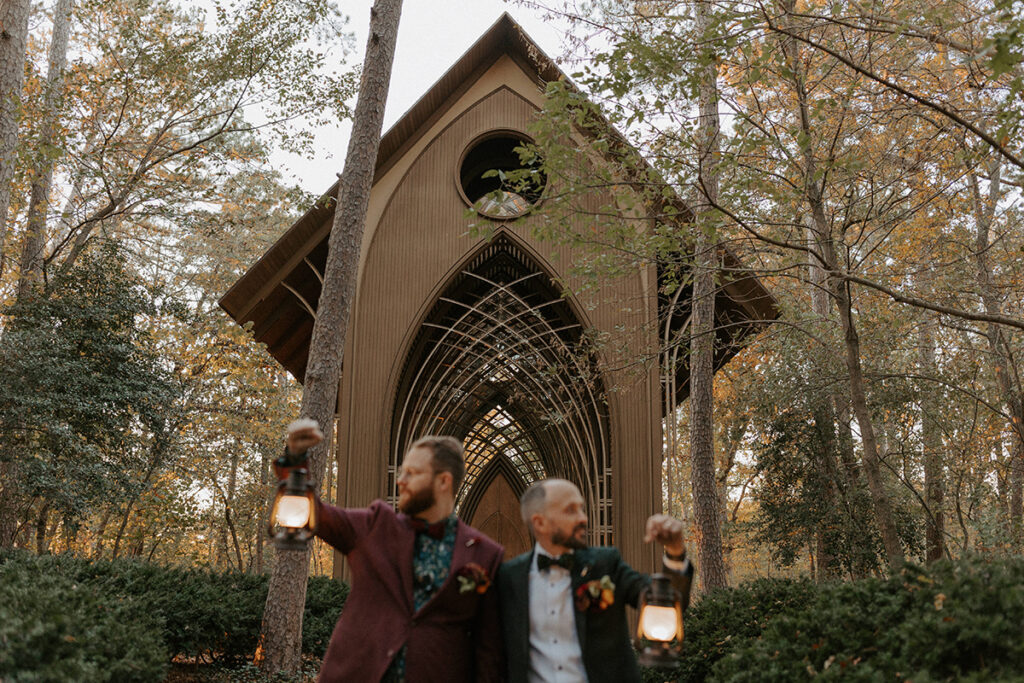 Two grooms in wedding attire hold lanterns as they look at the surrounding scenery of their wedding venue, Cooper Chapel in Bella Vista, Arkansas