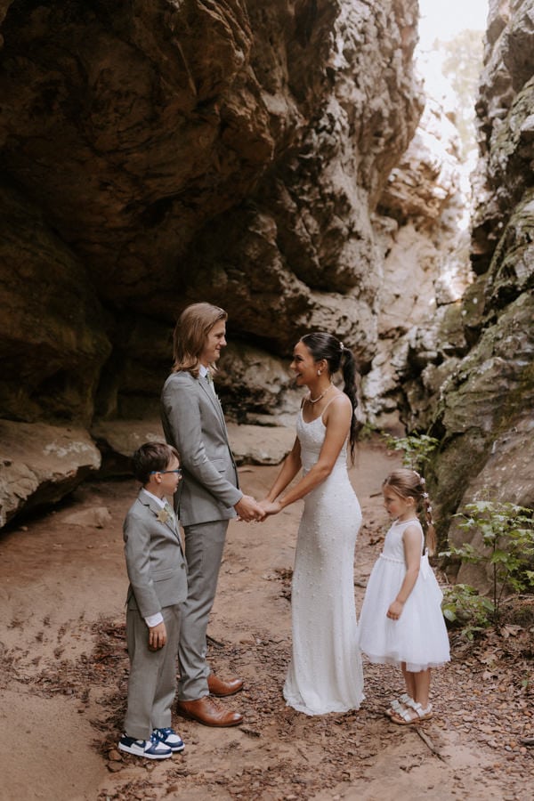 A bride and groom hold hands smiling and laughing with their two young children by each of their sides during their ceremony at Bear Cave Trail for their Arkansas elopement.