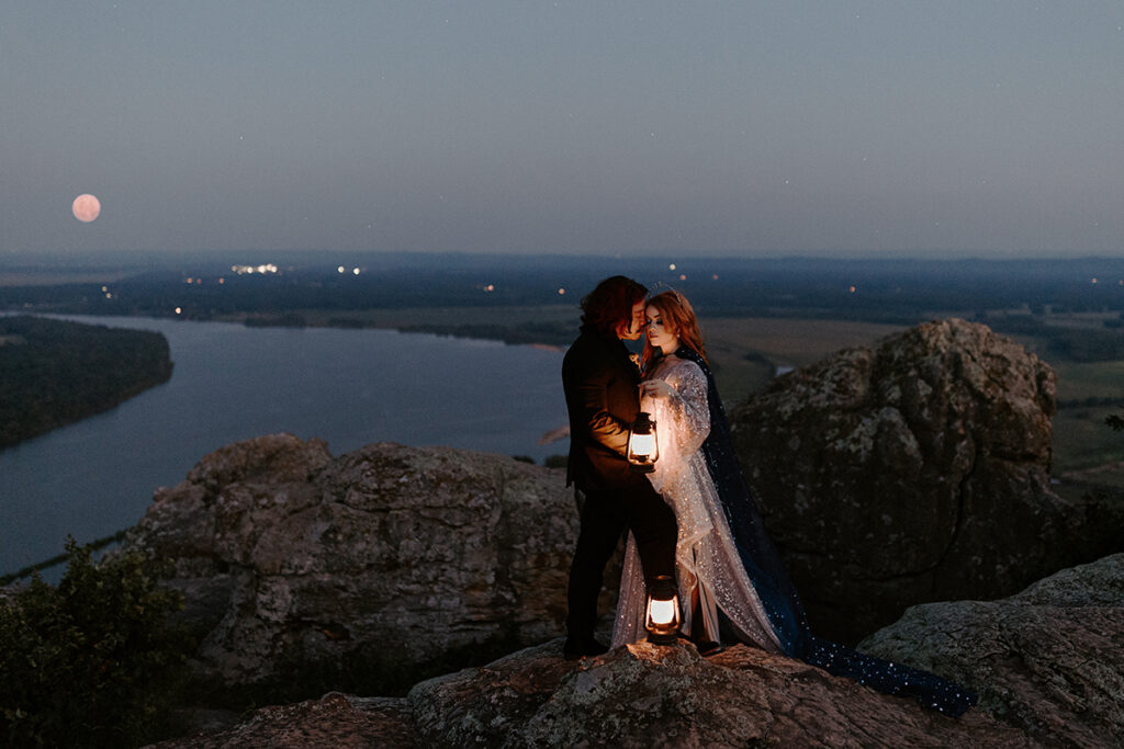 A bride and groom in wedding attire stand on the cliffside of Petit Jean State Park as the moon rises over the Arkansas River during their blue hour elopement.