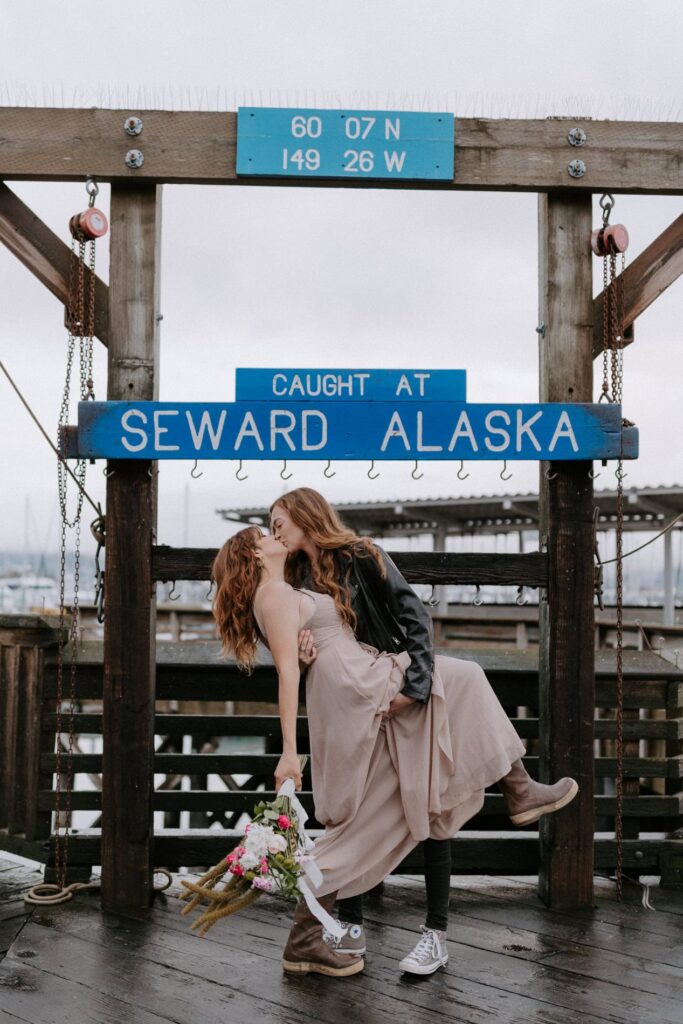 A bride dips down her spouse for a kiss in front of the "Caught at Seward Alaska" sign during their Kenai Fjords elopement.