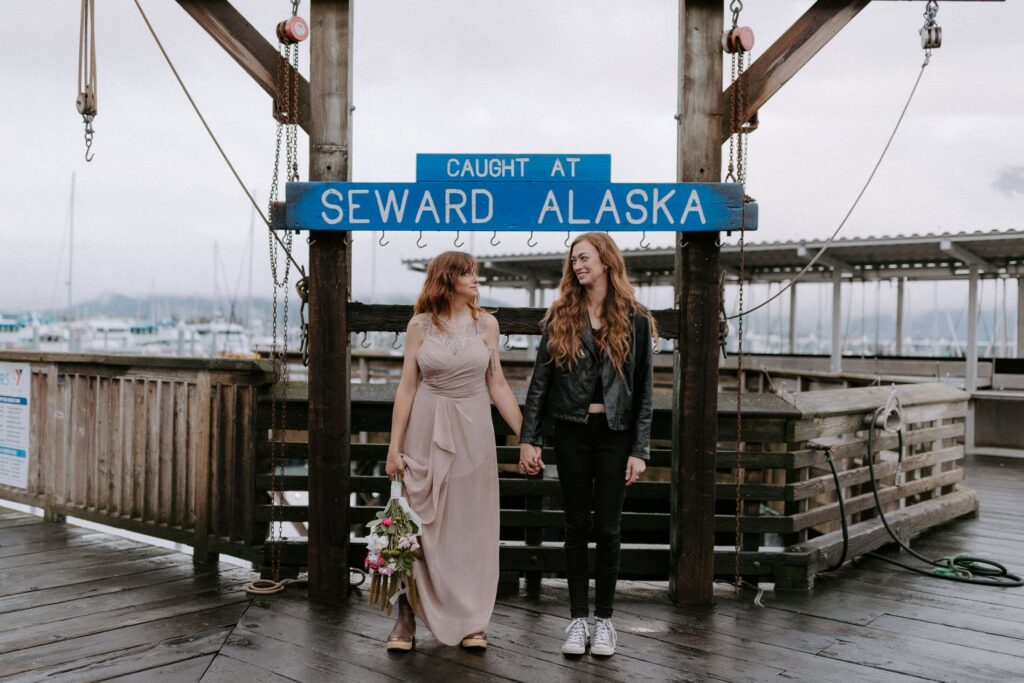 A couple looks at each other smiling in front of the "Caught at Seward Alaska" sign during their adventure elopement in the Kenai Fjords National Park.