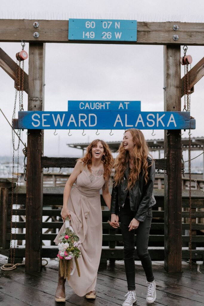 A couple laughing while trying to pose for an elopement photo in front of the "caught at Seward Alasak" sign at the Seward Boat Harbor.