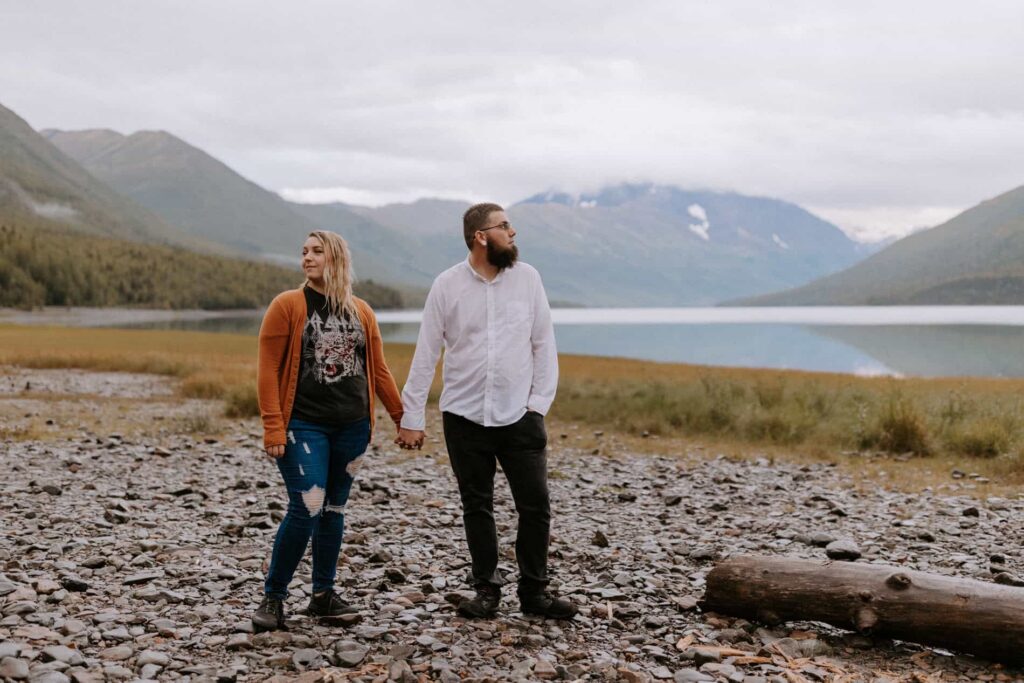 A couple holds hands posing on the lakeside of Eklunta Lake with scenic mountain views, known for being one of the best places to elope in Alaska.