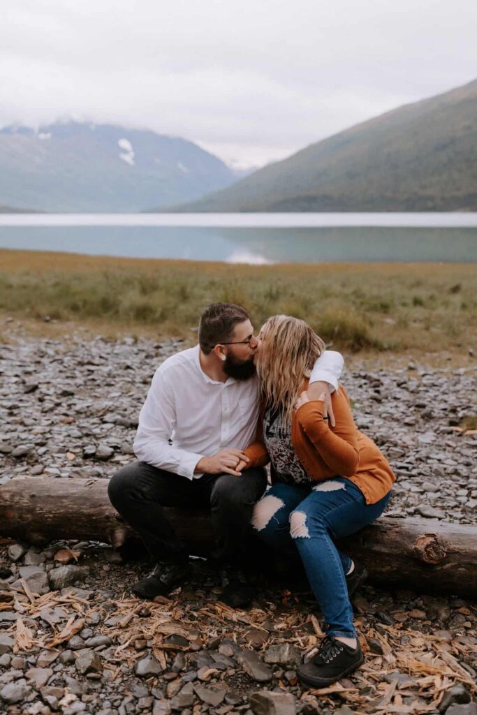 A couple sits on a fallen log kissing at Eklunta Lake, known for being one of the best places to elope in Alaska.