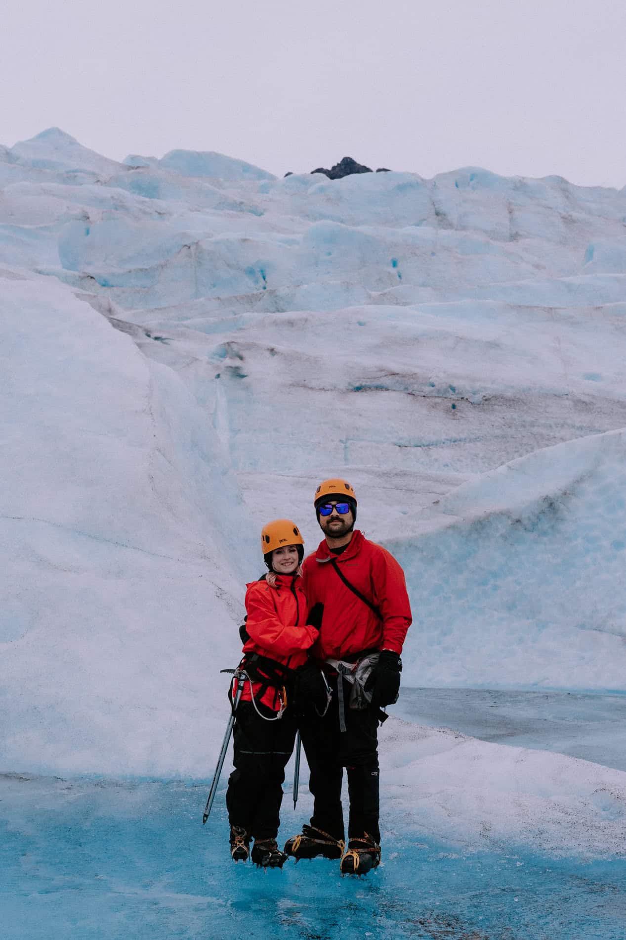 A couple poses for a photo in front of a small glacier waterfall while standing in a pool of very blue water on Mendenhall Glacier, one of the best places to elope in Alaska.