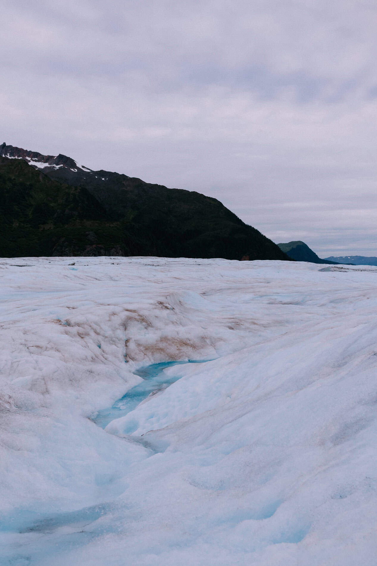 A glacial river flows across a wide icy landscape with tall mountains in the background on Mendenhall Glacier. One of the best places to elope in Alaska.