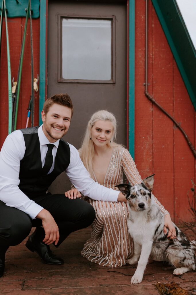 A bride and groom pet their dog as all three of them look at the camera for a photo during an adventure elopement at Hatcher Pass Lodge.