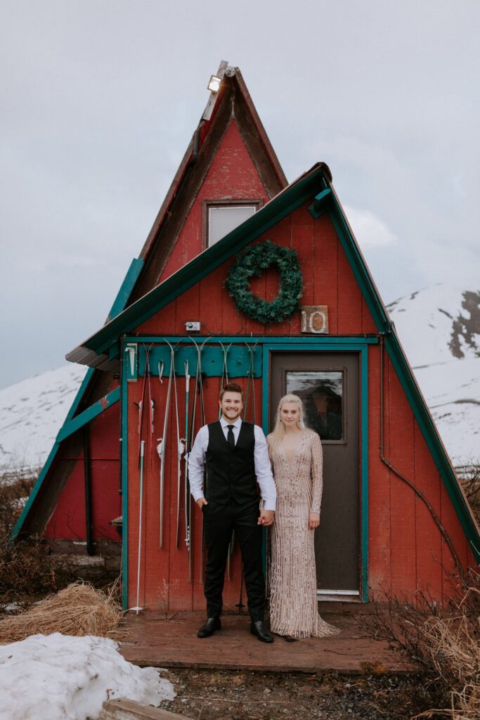 A couple stands in front of the iconic red cabins at Hatcher Pass during their wintry Alaska elopement.