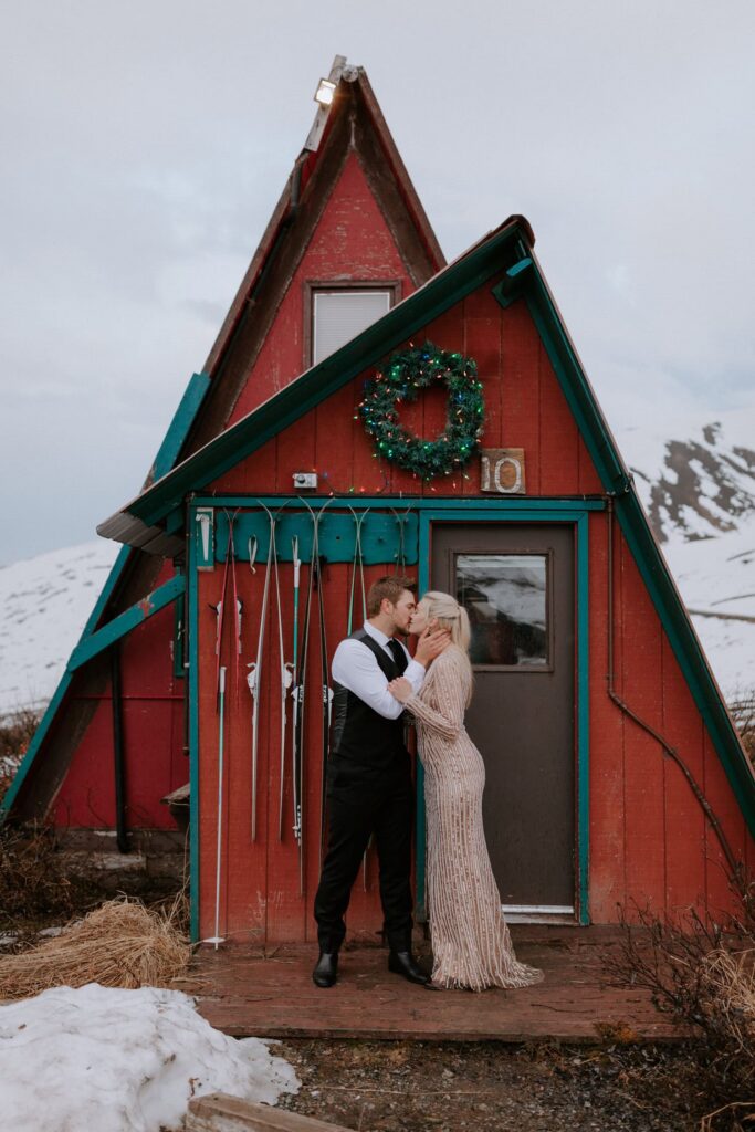 A couple kisses in front of the iconic red cabins at Hatcher Pass during an adventure elopement in Alaska.
