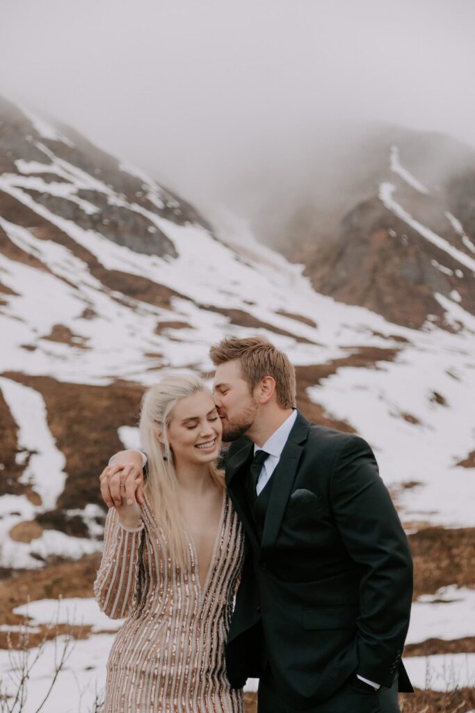 A groom kisses his bride on the cheek in front of a snowy mountain during their elopement at Hatcher Pass, Alaska.