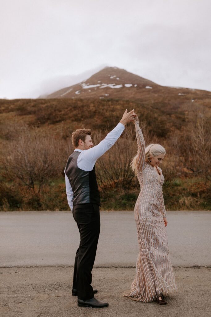 A groom twirls his bride with scenic views of a mountain in the background during their adventure elopement at Hatcher Pass in Alaska.