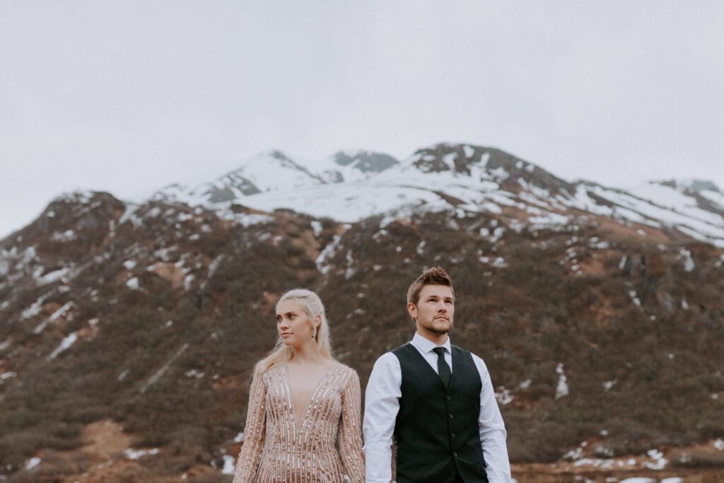 A couple stands side by side for a photo in front of a snow-capped mountain during their elopement at Hatcher Pass, Alaska.