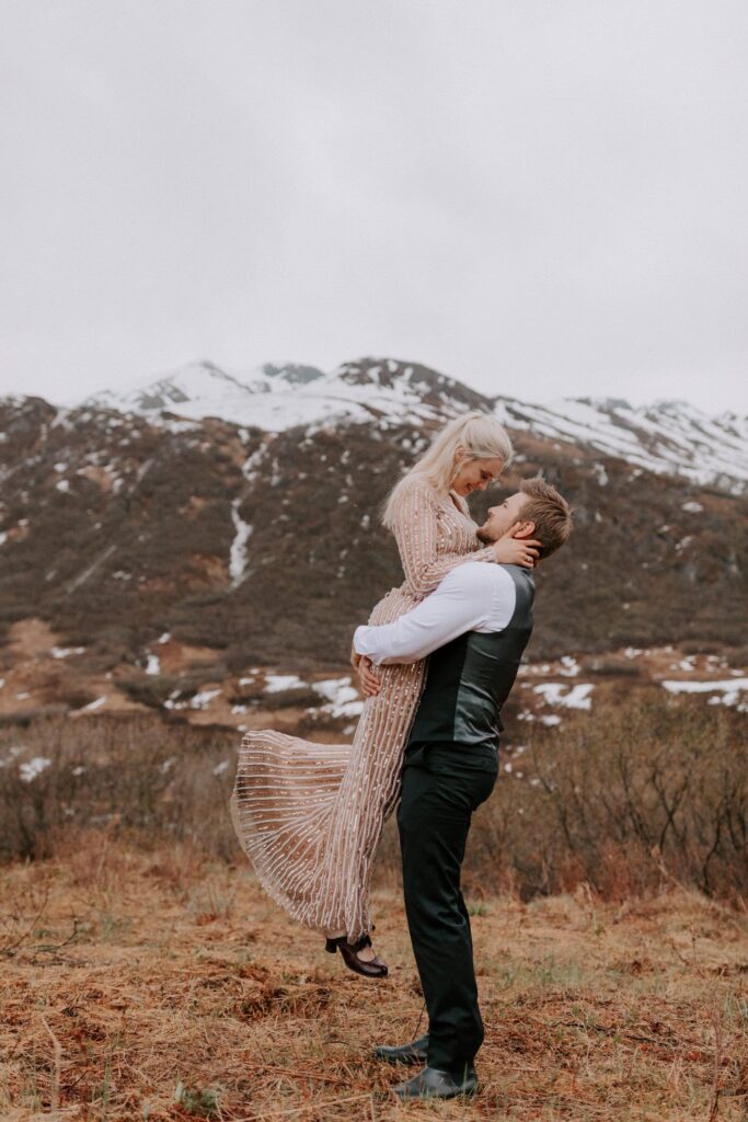 A groom picks up his bride in front of a snowy mountain during their Hatcher Pass elopement in Alaska.