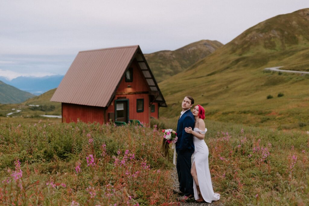 A wedding couple poses for a photo in front of the tiny red cabins at Hatcher Pass, Alaska.