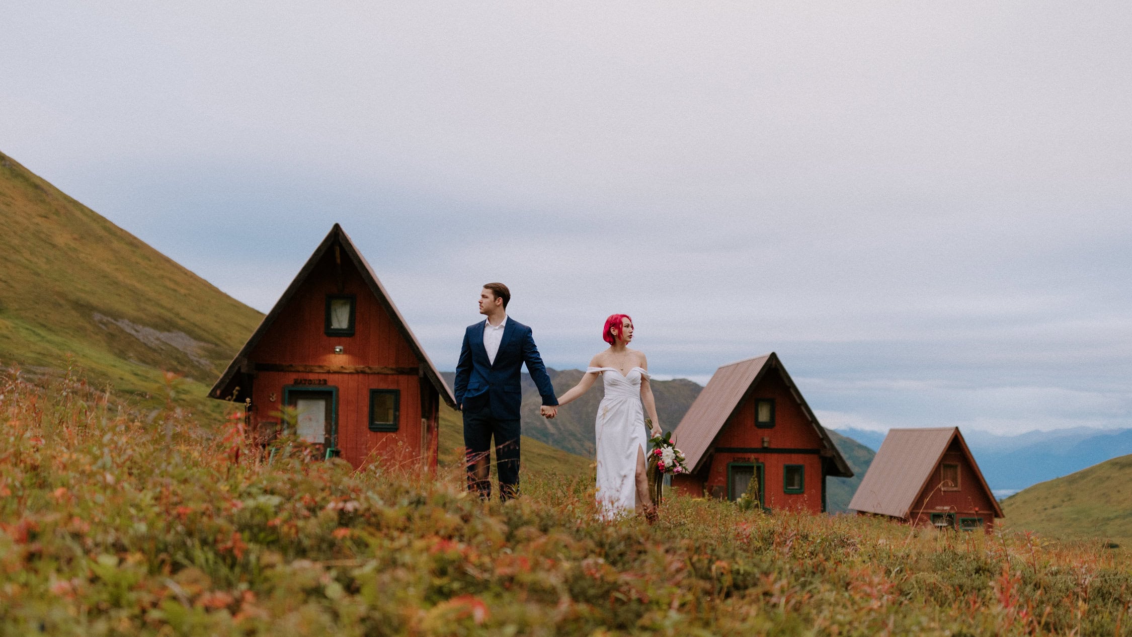 A bride and groom pose in front of the iconic tiny red cabins at Hatcher Pass for their Alaska elopement.