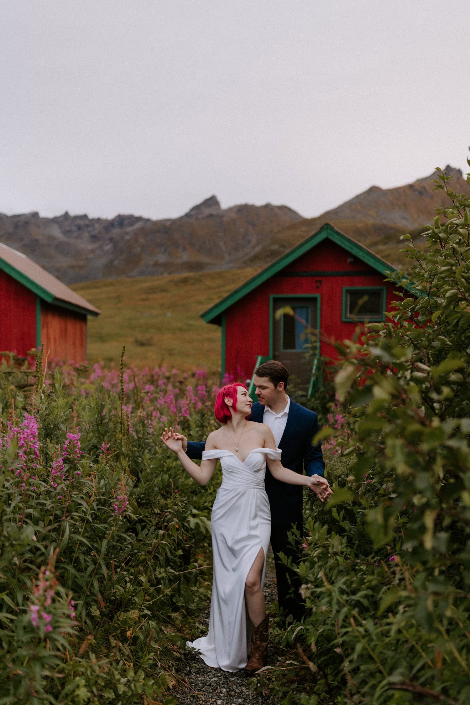 A bride and groom pose in front of a tiny red cabin during their Alaskan adventure elopement at Hatcher Pass.