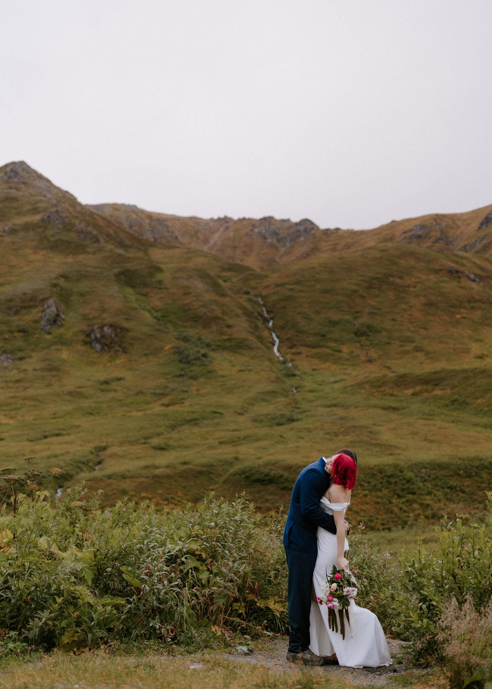 An elopement couple embraces in front of a small waterfall running down the mountains of Hatcher Pass.