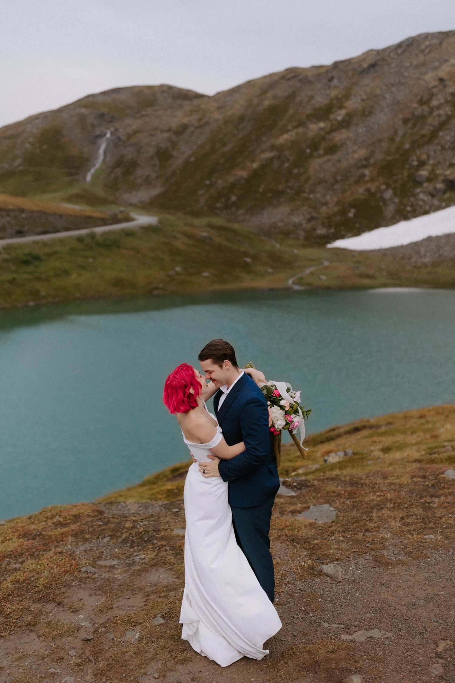 A couple embraces each other while standing in front of Summit Lake at Hatcher Pass during their Alaska adventure elopement.