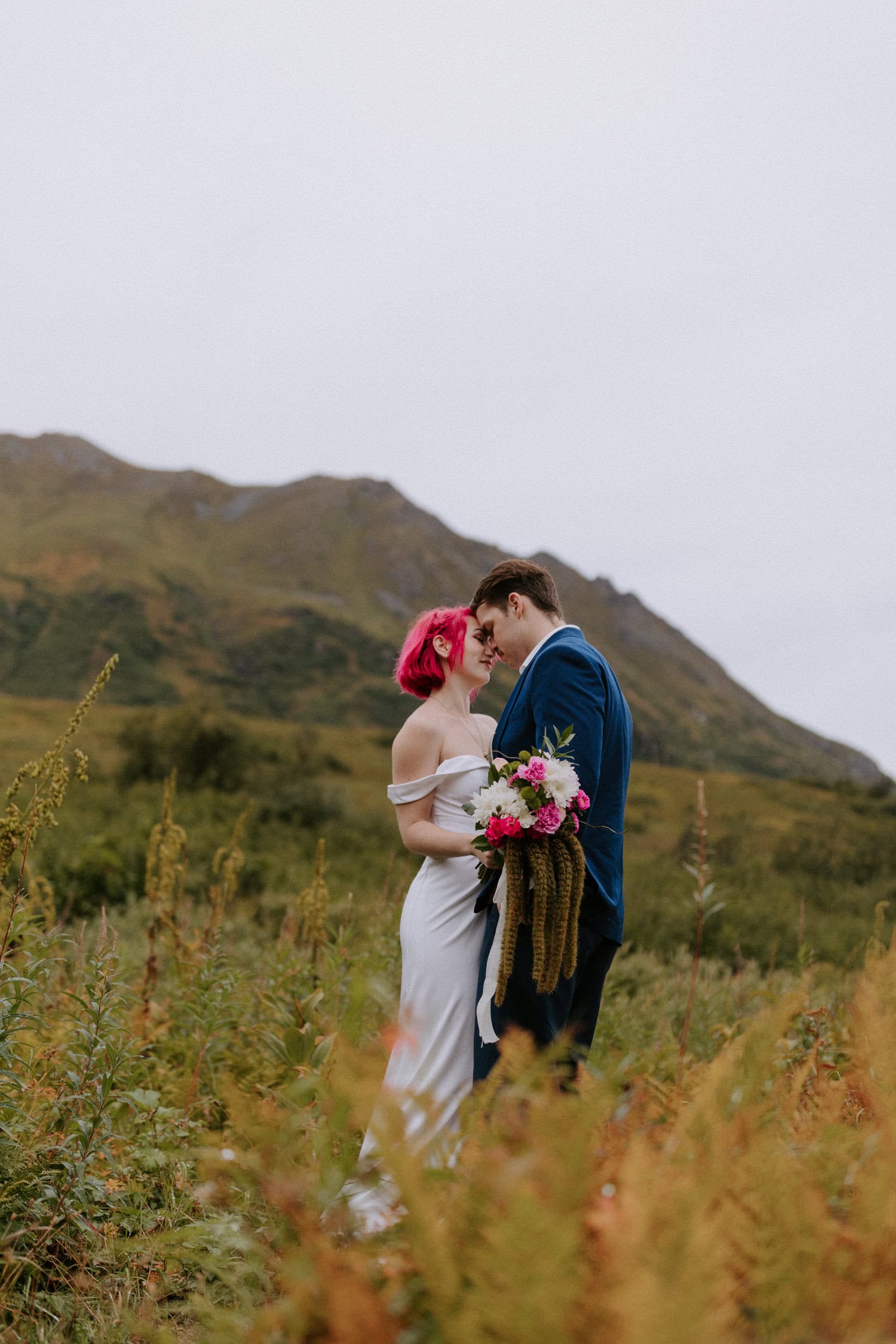 A couple stands closely together with their foreheads pressed while they pose for a photo in front of the mountains of Hatcher Pass.