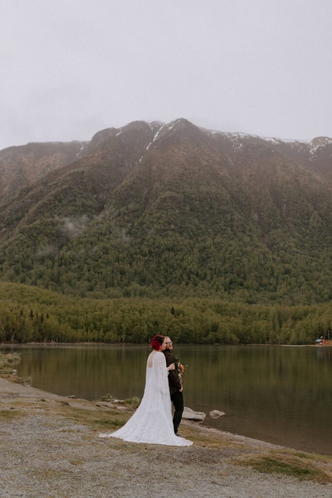 A couple embracing by the shoreline of Mirror Lake outside of Anchorage, Alaska with a tall misty mountain in the background.
